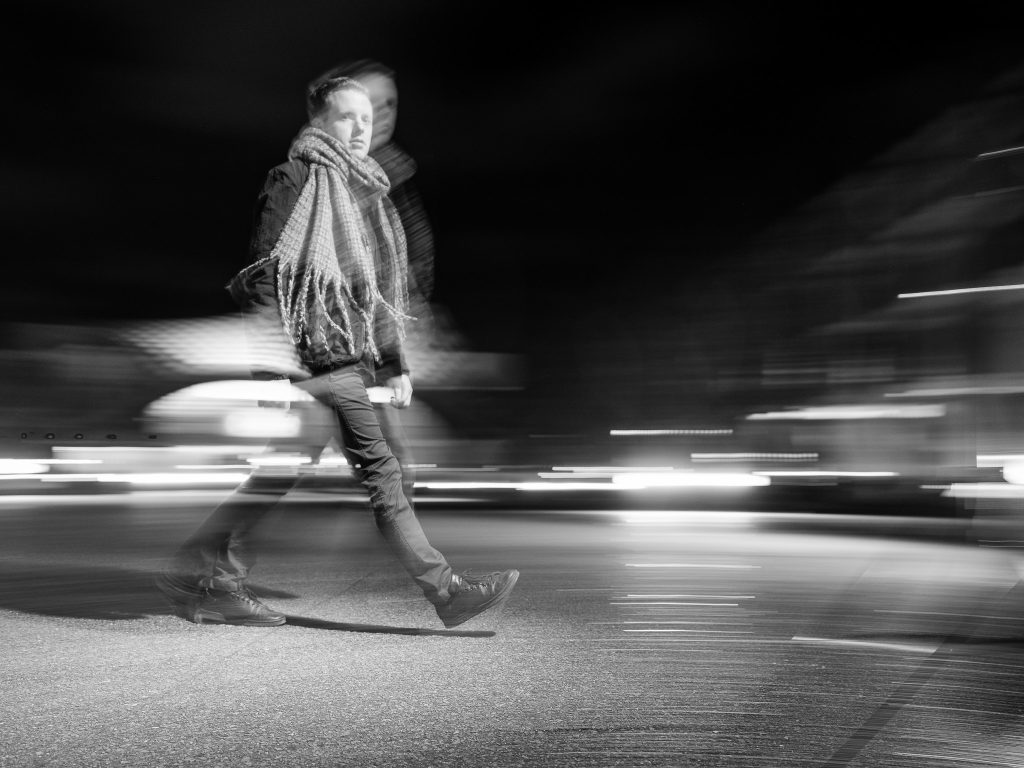 La solidarité des singularités : pourquoi j’aime mettre en avant ceux qu’on ne voit pas 2 Un homme en écharpe traverse la ville de nuit en noir et blanc, capturé dans un effet de filé, symbolisant le mouvement et l’individualité.