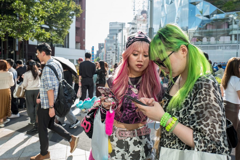 La solidarité des singularités : pourquoi j’aime mettre en avant ceux qu’on ne voit pas 1 Deux jeunes femmes aux styles colorés et originaux marchent dans une rue animée de Tokyo, illustrant la diversité et l’expression personnelle.