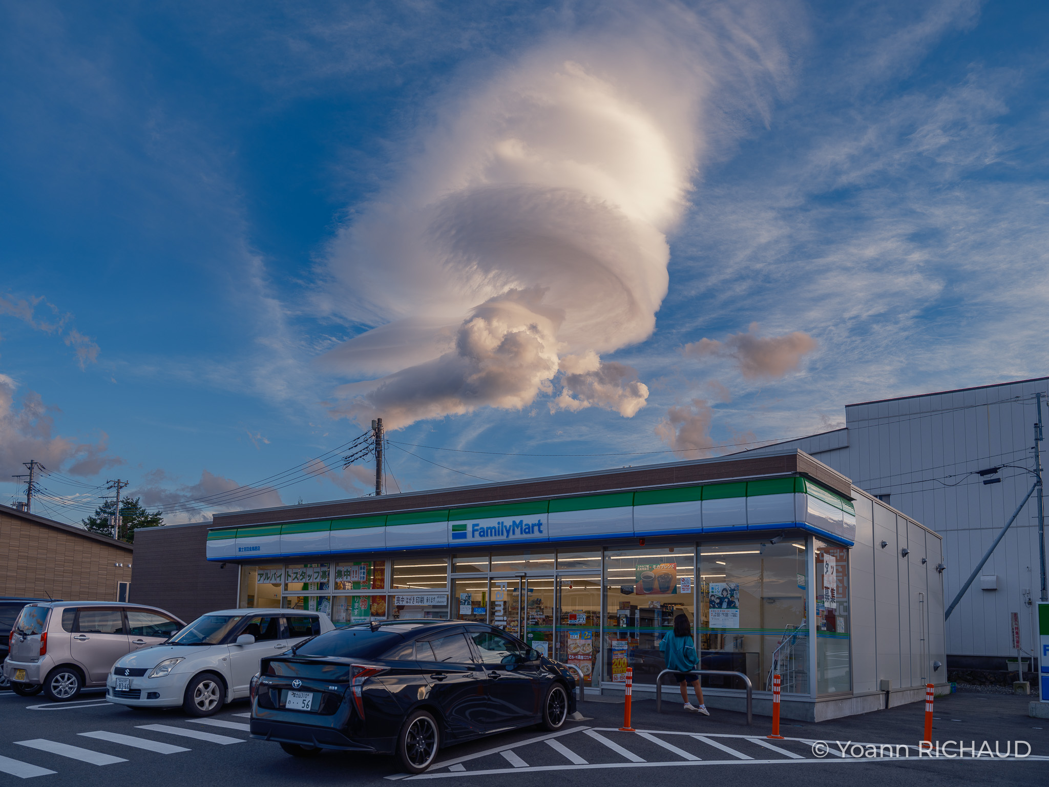 Supérette et voitures sous un ciel bleu traversé par un grand nuage clair.