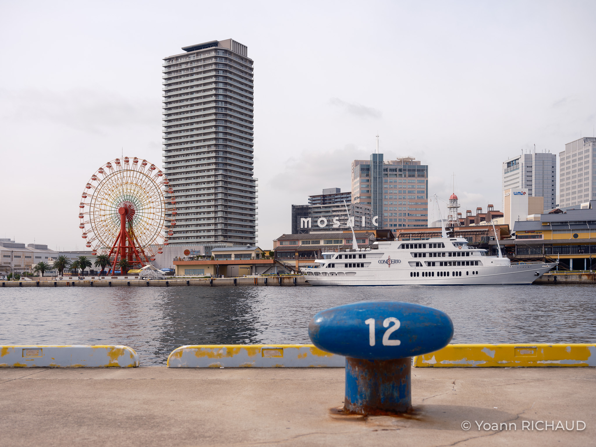 Grande roue rouge et immeubles au bord de l’eau, avec une borne bleue au premier plan.